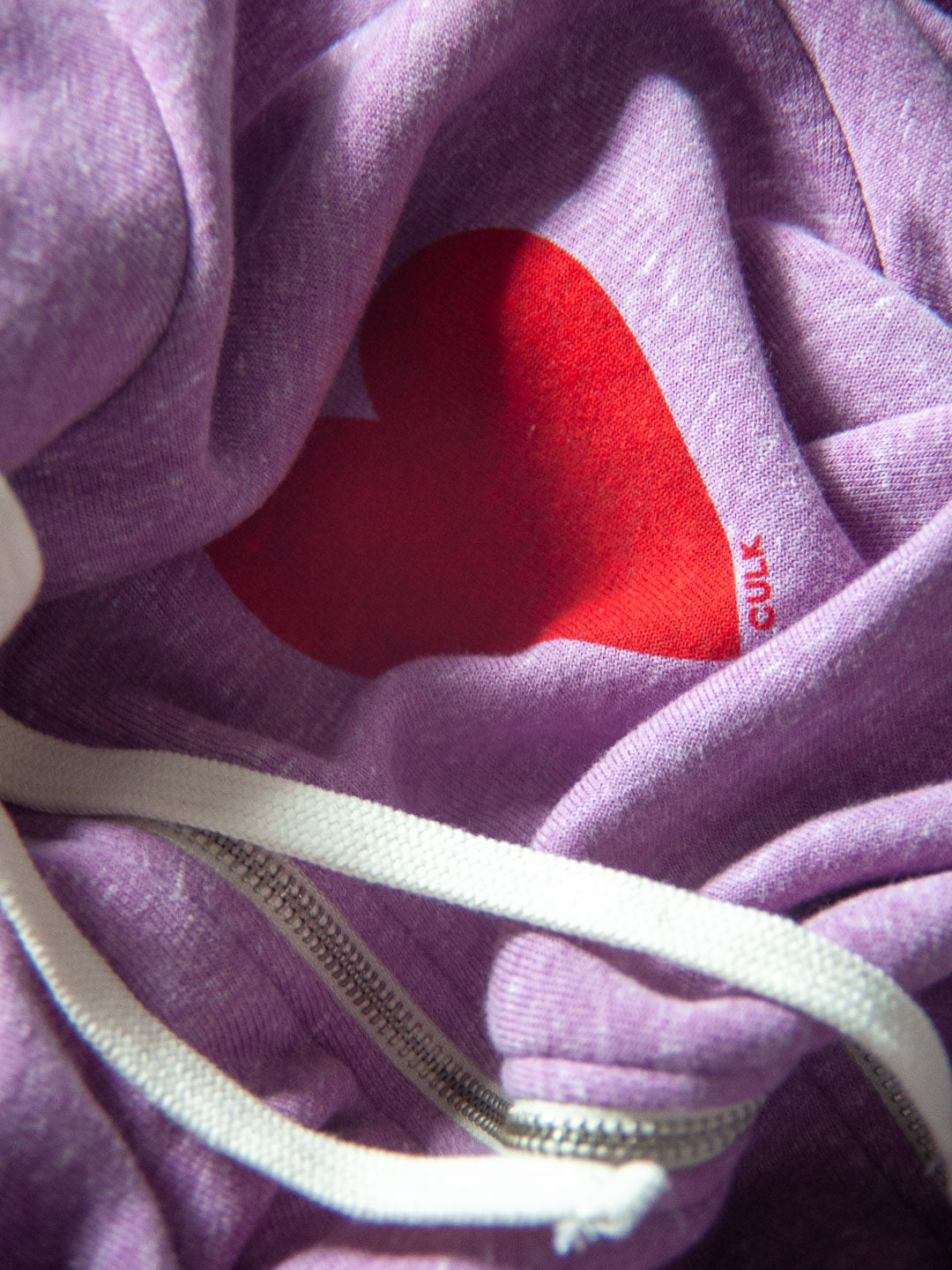 Close-up of a purple zip-up hoodie with a red heart-shaped patch on the inside collar and a white drawstring.