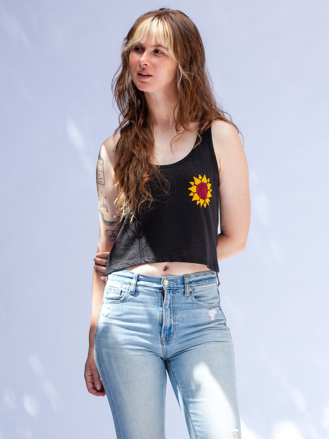 Woman wearing a black tank top with a sunflower design and light blue jeans against a light gray background