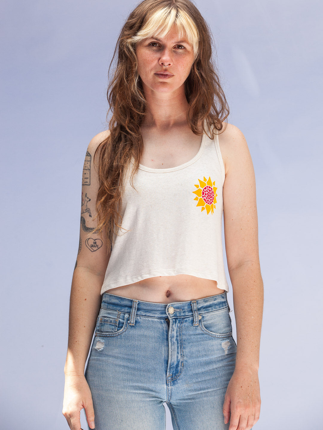 Person wearing a white tank top with a colorful flower design and blue jeans against a light gray background